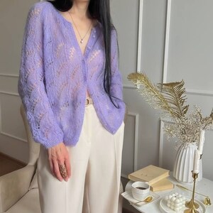 May include: A light purple, open-weave cardigan with a button-up front. The cardigan is paired with cream-colored wide-leg pants. The outfit is completed with a gold ring and a delicate necklace. The cardigan's sleeves are long and the fabric appears soft.