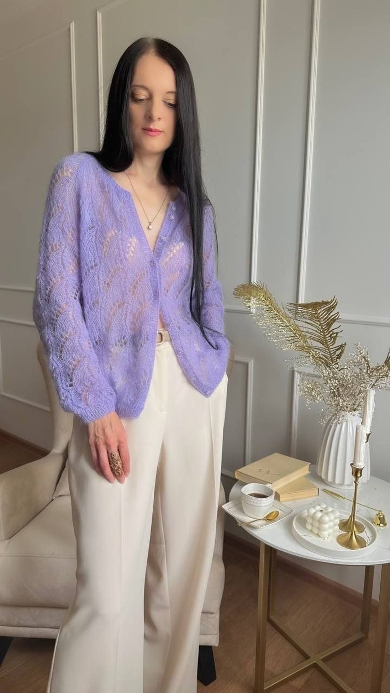 May include: A light purple, open-weave cardigan with a button-up front. The cardigan is paired with cream-colored wide-leg pants. The outfit is completed with a gold ring and a delicate necklace. The cardigan's sleeves are long and the fabric appears soft.