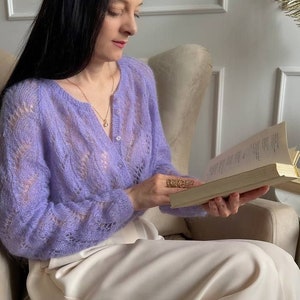 May include: A light purple, open-weave cardigan with a V-neck and button closure. The cardigan is paired with cream-colored pants. The woman is wearing a gold necklace and ring while reading a book. The setting is a neutral-toned room.