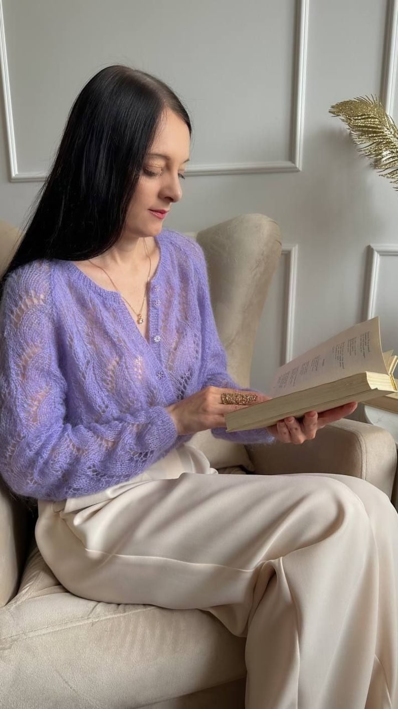 May include: A light purple, open-weave cardigan with a V-neck and button closure. The cardigan is paired with cream-colored pants. The woman is wearing a gold necklace and ring while reading a book. The setting is a neutral-toned room.