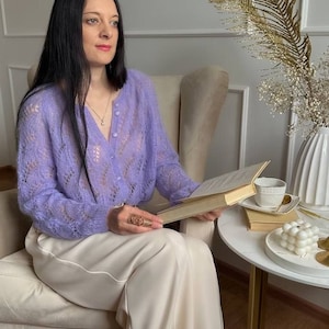 May include: A woman wearing a light purple, open-knit cardigan and cream-colored pants, seated in a beige armchair, holding a book. A small white table holds a cup, saucer, and decorative items. The background features a white vase with gold accents.