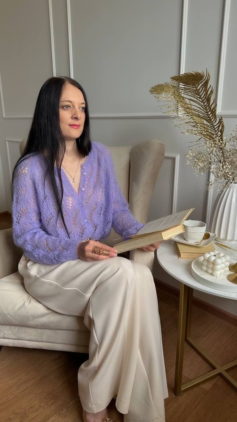 May include: A woman wearing a light purple, open-knit cardigan and cream-colored pants, seated in a beige armchair, holding a book. A small white table holds a cup, saucer, and decorative items. The background features a white vase with gold accents.
