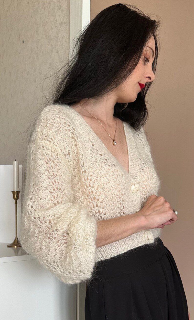 May include: Cream-colored, open-knit cardigan with a V-neck and button closure. The long-sleeved sweater has a delicate, lacy pattern and is paired with a black skirt. A gold necklace with a pendant is visible.