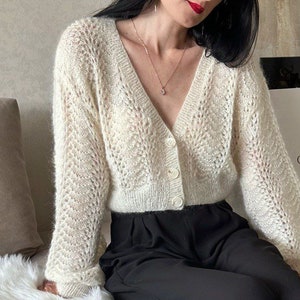 May include: Cream-colored, open-weave cardigan with a V-neck and button closure. The long-sleeved sweater has a delicate, lacy pattern. The model is wearing black pants and a gold necklace with a pendant.