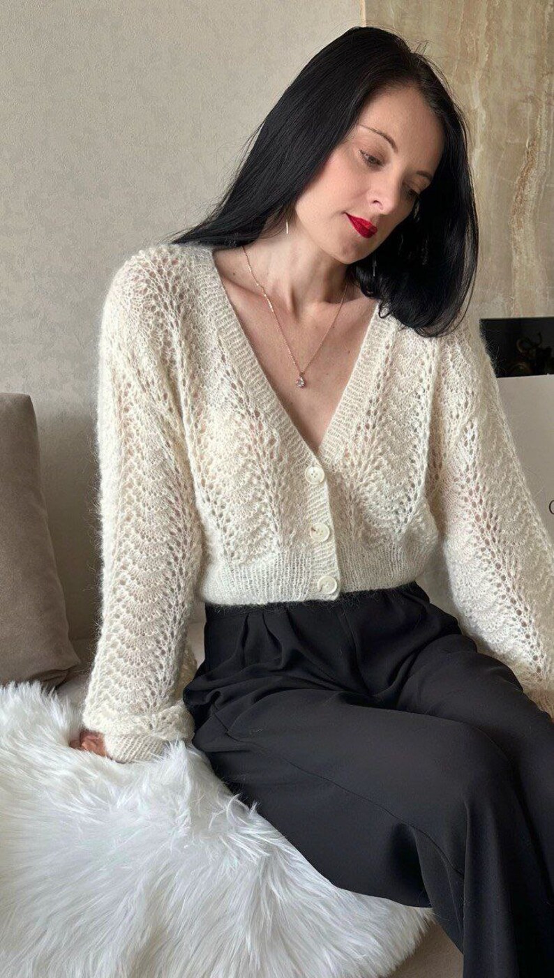 May include: Cream-colored, open-weave cardigan with a V-neck and button closure. The long-sleeved sweater has a delicate, lacy pattern. The model is wearing black pants and a gold necklace with a pendant.