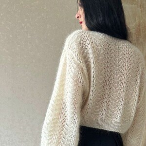 May include: Cream-colored, open-knit sweater with long, voluminous sleeves. The sweater has a delicate, textured pattern and is paired with a black skirt. The image is a back view, showcasing the sweater's design and fit.