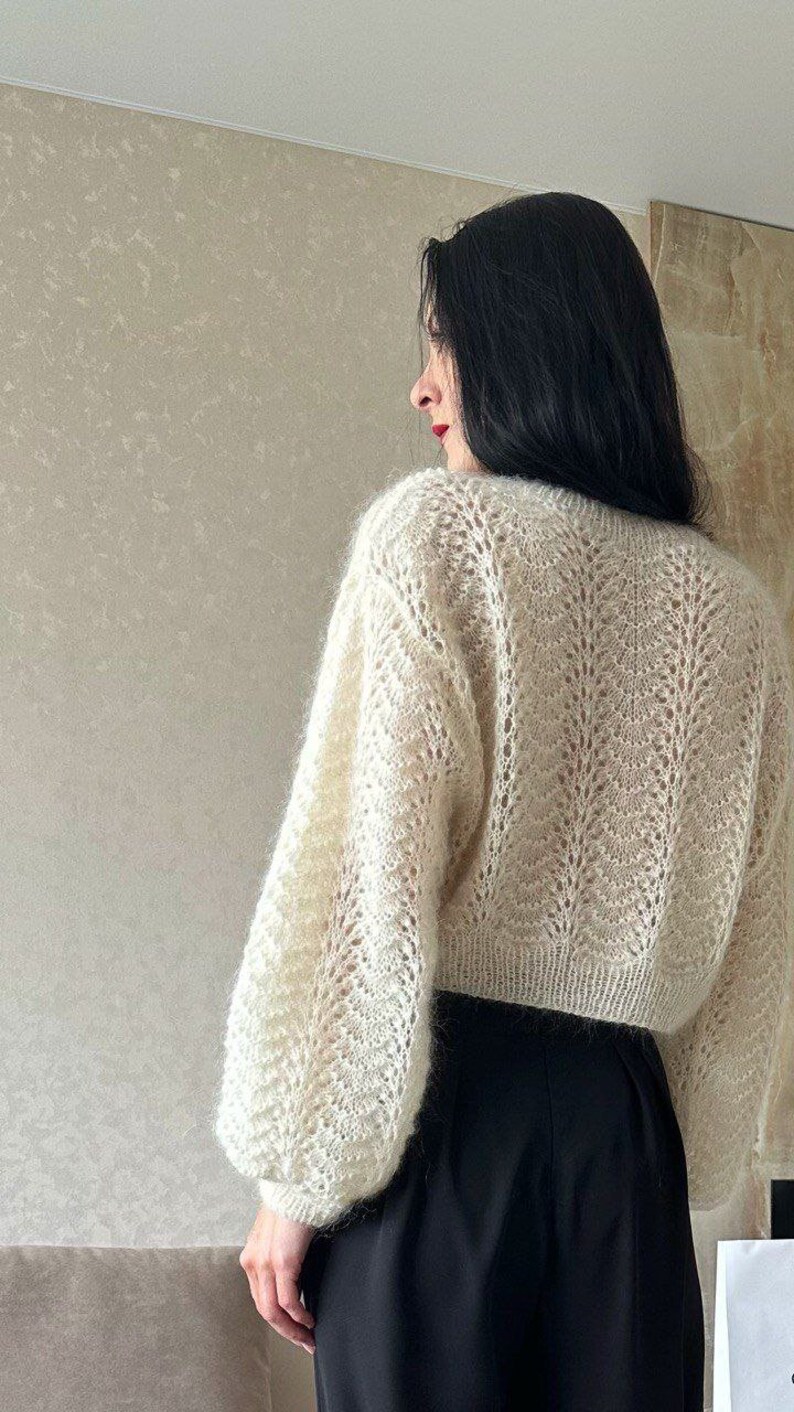 May include: Cream-colored, open-knit sweater with long, voluminous sleeves. The sweater has a delicate, textured pattern and is paired with a black skirt. The image is a back view, showcasing the sweater's design and fit.