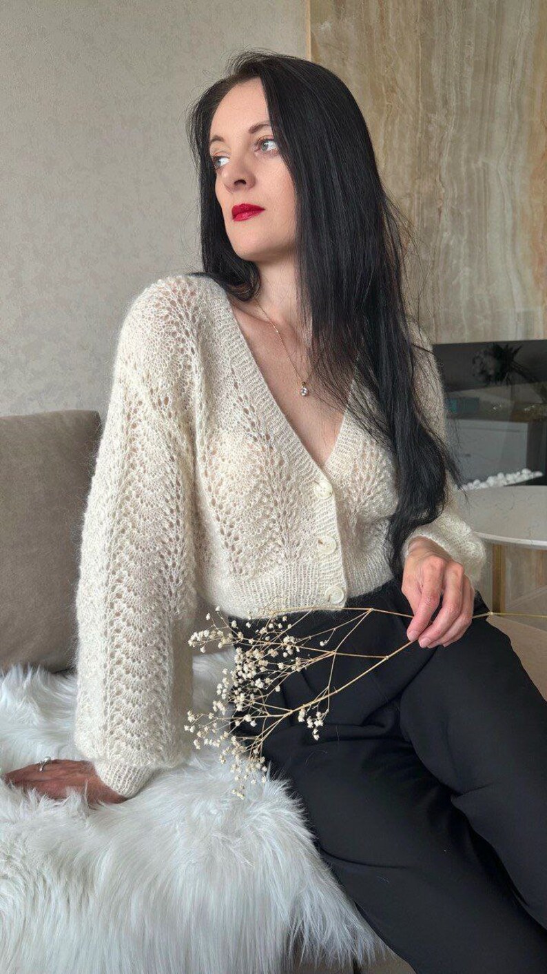 May include: Cream-colored, open-weave cardigan with a V-neck and button closure. The long-sleeved sweater has a delicate, lacy pattern. The model is wearing black pants and a necklace. The cardigan is a stylish addition to any wardrobe.