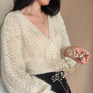 May include: Cream-colored, open-knit cardigan with a V-neck and three pearl-like buttons. The long-sleeved sweater has a delicate, lacy pattern. The woman is wearing a gold necklace and holding a sprig of white flowers. The woman is wearing black pants.