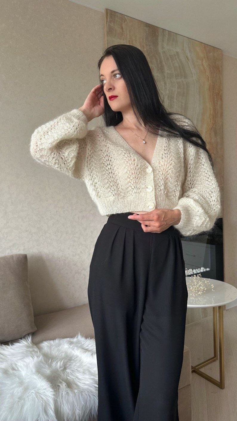 May include: Cream-colored, open-knit cardigan with a V-neck and button closure. The cropped sweater is paired with high-waisted, black trousers. The outfit is completed with a delicate necklace. The overall style is casual and elegant.