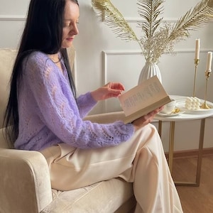 May include: A person seated in a beige armchair, reading a book. They are wearing a light purple, open-knit sweater and cream-colored pants. Gold-colored heels with decorative embellishments are visible. A small side table holds candles and a vase with decorative foliage.