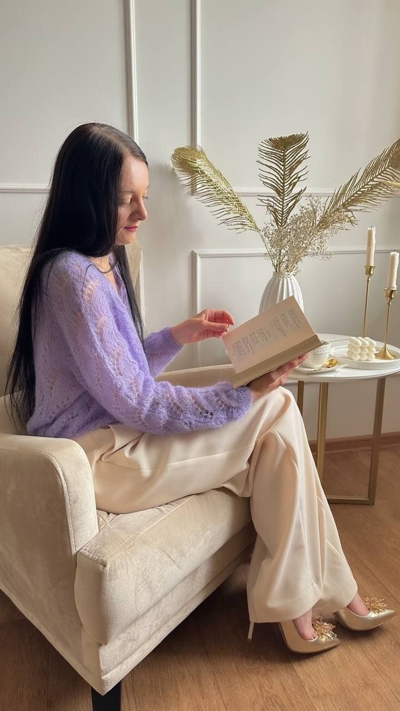 May include: A person seated in a beige armchair, reading a book. They are wearing a light purple, open-knit sweater and cream-colored pants. Gold-colored heels with decorative embellishments are visible. A small side table holds candles and a vase with decorative foliage.