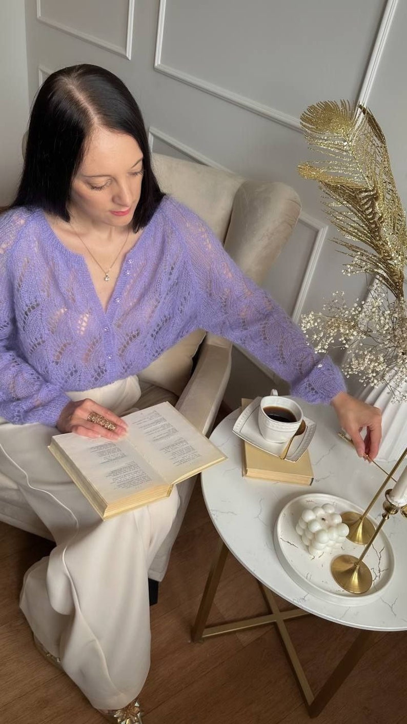 May include: A person wearing a light purple, open-knit cardigan, reading a book. A small white table holds a cup of coffee, a decorative plate, and a candle holder. The room has a neutral color palette.