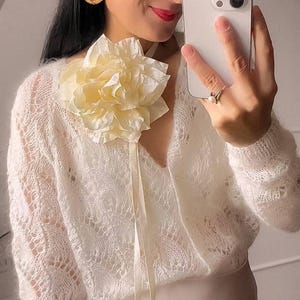 May include: A woman wearing a white, open-knit sweater with a large, cream-colored flower pinned to the neckline. The sweater has long sleeves and a V-neck. The woman is holding a smartphone and wearing gold earrings.