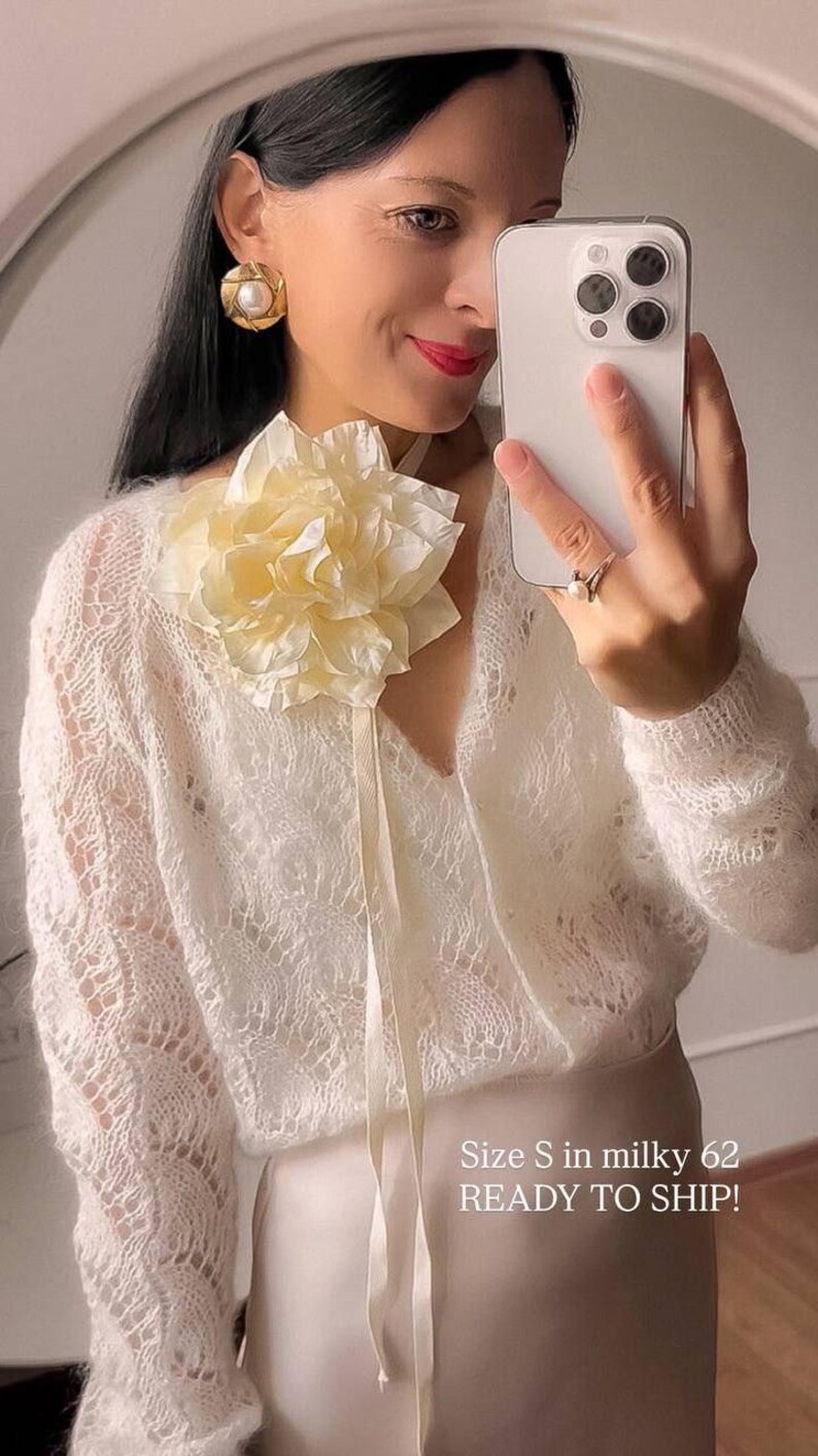 May include: A woman wearing a white, open-knit sweater with a large, cream-colored flower pinned to the neckline. The sweater has long sleeves and a V-neck. The woman is holding a smartphone and wearing gold earrings.