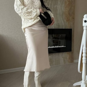 Op de afbeelding: Een ivoorkleurige, open gebreide trui met lange mouwen wordt gecombineerd met een lichtroze satijnen midi-rok en witte kniehoge laarzen. Een zwarte clutch en een cr&egrave;mekleurige fedora maken de outfit compleet. De achtergrond toont een open haard en een decoratieve muur.