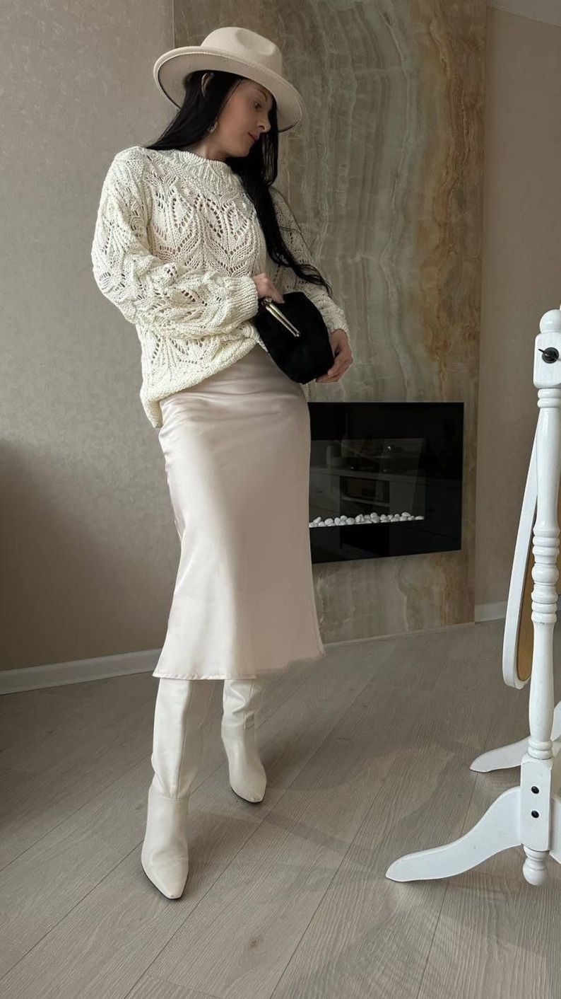Op de afbeelding: Een ivoorkleurige, open gebreide trui met lange mouwen wordt gecombineerd met een lichtroze satijnen midi-rok en witte kniehoge laarzen. Een zwarte clutch en een cr&egrave;mekleurige fedora maken de outfit compleet. De achtergrond toont een open haard en een decoratieve muur.
