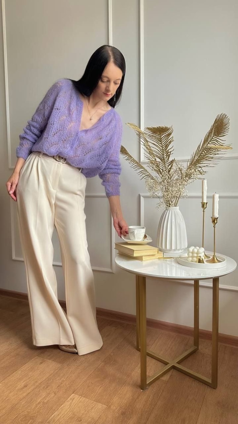 May include: A woman wearing a light purple, open-knit cardigan and cream-colored wide-leg pants. A small, round table with a white marble top holds a white vase with dried flowers, candles, and a teacup. The table has gold legs.