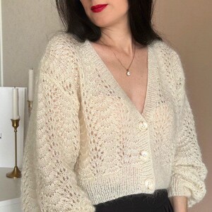 May include: Cream-colored, open-weave cardigan with a V-neck and three large, round buttons. The sleeves are long and slightly puffed. The cardigan is paired with black pants. A gold necklace is visible.