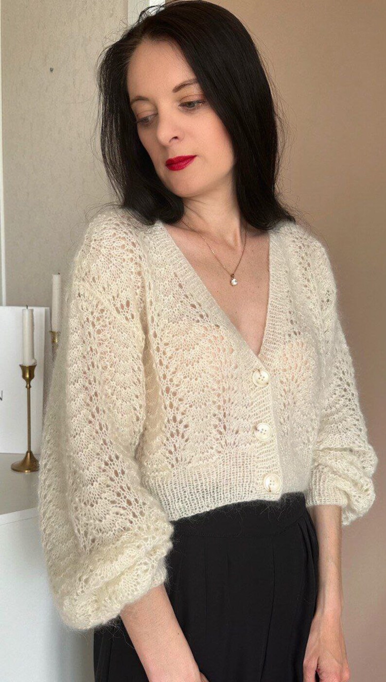 May include: Cream-colored, open-weave cardigan with a V-neck and three large, round buttons. The sleeves are long and slightly puffed. The cardigan is paired with black pants. A gold necklace is visible.