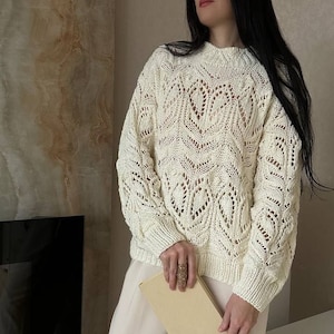 Op de afbeelding: Cr&egrave;mekleurige, open gebreide trui met een ronde hals en lange mouwen. De trui heeft een gedetailleerd, patroonontwerp. De persoon draagt een cr&egrave;mekleurige broek en houdt een boek vast. De achtergrond omvat een open haard en een neutraal getinte muur.