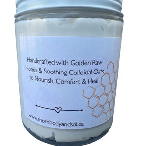 May include: A glass jar with a white lid, containing a creamy product. The label on the jar states "Handcrafted with Golden Raw Honey & Soothing Colloidal Oats to Nourish, Comfort & Heal." The website www.mombodyandsol.ca is also visible.