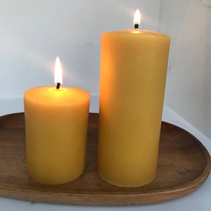 May include: Two beeswax candles, one larger than the other, are lit and burning on a wooden tray. The candles are a pale yellow colour.