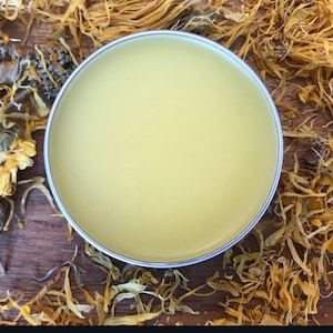 May include: A circular, silver-edged container holding a smooth, pale yellow balm. The balm is set against a backdrop of dried calendula flowers and petals in shades of yellow and brown. This suggests a natural skincare product.