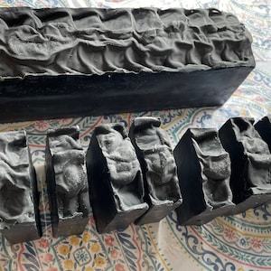 May include: A large black bar of soap and several smaller bars of black soap. The soap has a textured surface.