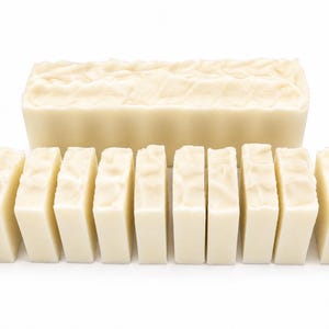 May include: A selection of handmade, off-white soap bars. A large rectangular bar with a textured top is at the back. In front are several smaller, rectangular bars, also off-white, cut from the larger bar. The soaps are arranged on a white background.