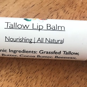 May include: White lip balm tube with the text "Tallow Lip Balm" and "Nourishing | All Natural". The label lists organic ingredients: Grassfed Tallow, Shea Butter, Cocoa Butter, and Beeswax. The tube is on a wooden surface.