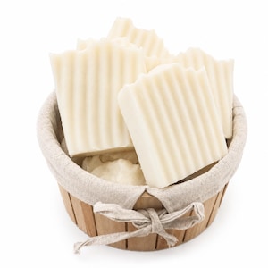 May include: A wooden basket containing several bars of off-white soap. The soap bars have a ridged texture and are rectangular. The basket has a linen lining and a decorative bow. The soap is likely handmade and natural.