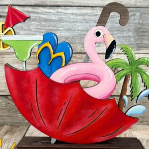 May include: A wooden flamingo pool float, flip-flops, and a margarita are under a red umbrella. A palm tree and water splashes are also included. The base has the text "CUTSUN" in a yellow sun.