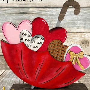 May include: A wooden Valentine's Day decoration featuring a red umbrella shape holding heart-shaped cutouts. The hearts are pink, red, and white, with waffle and gift box details. The text "digital download" and "CUTSUN" are visible.