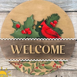 以下が含まれることがあります： 上部にCHRISTMAS ADD-ON、中央にWELCOMEの文字が記された円形の木製看板。赤いショウジョウコウカンチョウ、赤い実をつけた緑のヒイラギの葉、そして下部には葉と実の植物模様がデザインされています。