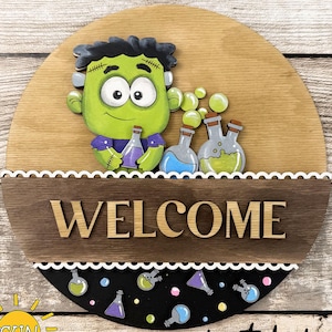 May include: A Halloween-themed wooden sign with a cartoon Frankenstein's monster holding potion bottles. The sign features the text "WELCOME" and "HALLOWEEN ADD-ON". The design includes a black and white scalloped border and a digital download graphic.