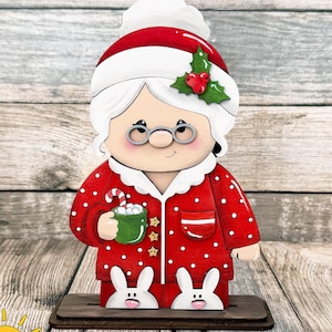 May include: A wooden cutout of a grandmother figure wearing a red and white polka dot pajama set, a matching nightcap with holly, and white bunny slippers. The figure holds a green mug with marshmallows and a candy cane, wearing round glasses.