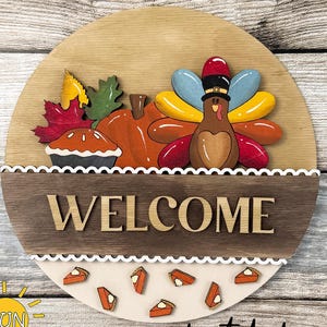 以下が含まれることがあります： 上部にTHANKSGIVING ADD-ON、中央にWELCOMEの文字が書かれた円形の木製看板。七面鳥、カボチャ、パイ、秋の葉のデザインが施されています。下部には小さなパイのピースが配置され、digital downloadの文字が添えられています。