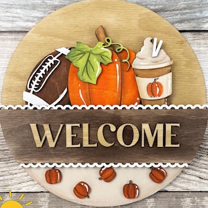 May include: A round wooden sign with a fall theme. It features a football, an orange pumpkin, and a coffee cup with a pumpkin design. The word "WELCOME" is displayed on a wooden banner. Small pumpkins and the text "Digital Download" are also present.