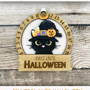 May include: A black cat wearing a witch's hat with candy corn and a pumpkin on top. The cat is in front of a wooden circle with the numbers 1 through 19. The circle says "Days Until Halloween".