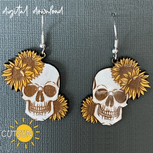 May include: A pair of wooden earrings featuring a white skull with brown details and a yellow sunflower design. The earrings are on a gray background.