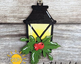 May include: A wooden Christmas lantern ornament with a black top and a yellow light. The ornament is decorated with green holly leaves and red berries. The text "CUTSUN" and "digital download" are visible.