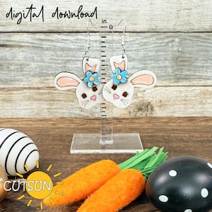 May include: White bunny rabbit earrings with blue flower accents. The earrings are hanging from a silver hook and are displayed on a clear acrylic stand. The background is a wooden surface.