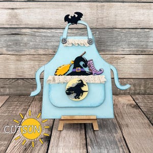 May include: A light blue apron-shaped decoration with a witch theme. It features a black bat, witch hat, broom, and boot. The apron has a pocket with a witch silhouette and the text "CUTSUN" in a sun graphic.