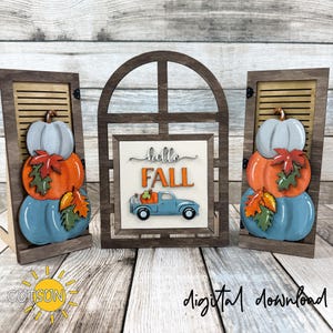 Könnte beinhalten: Holzdekor-Set zum Thema Herbst. Ein zentraler bogenförmiger Rahmen zeigt "hello FALL" und einen blauen Truck mit Kürbissen. Zwei Holzstücke im Fensterladen-Stil auf jeder Seite zeigen Stapel von blauen, orangefarbenen und grauen Kürbissen mit Herbstblättern.
