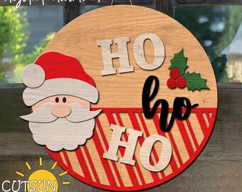 May include: A round wooden door hanger with a red and white striped pattern. The top half of the hanger features the words "HO HO" in white lettering. The bottom half features the words "HO HO" in black lettering. A Santa Claus face with a red hat is on the left side of the hanger. A green holly sprig with red berries is on the right side of the hanger.