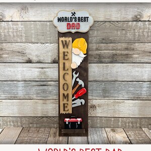 May include: Interchangeable porch sign with the text "World's Best Dad" and a gnome holding a welcome sign. The sign is decorated with tools and a toolbox. The sign is made for laser cutters.