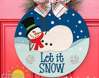 May include: A round wooden sign with a blue background and a snowman design. The snowman is white with black eyes and a carrot nose. The sign says "Let it Snow" in white letters. The sign is decorated with snowflakes and a red and green plaid bow.