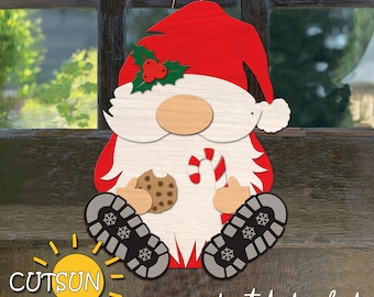Puede incluir: Un gnomo navideño rojo y blanco que sostiene un bastón de caramelo y una galleta de chocolate. El gnomo lleva un gorro rojo con una rama de acebo verde y una barba blanca. El gnomo está sentado con las piernas cruzadas y sus pies son negros con copos de nieve blancos.