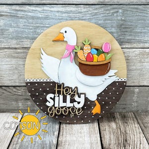 Könnte beinhalten: Runder Holzschild mit einer weißen Gans, die einen rosa Schal trägt und einen Korb mit Ostereiern und Karotten trägt. Auf dem Schild steht "Hey Silly Goose" und ein Sonnenlogo.